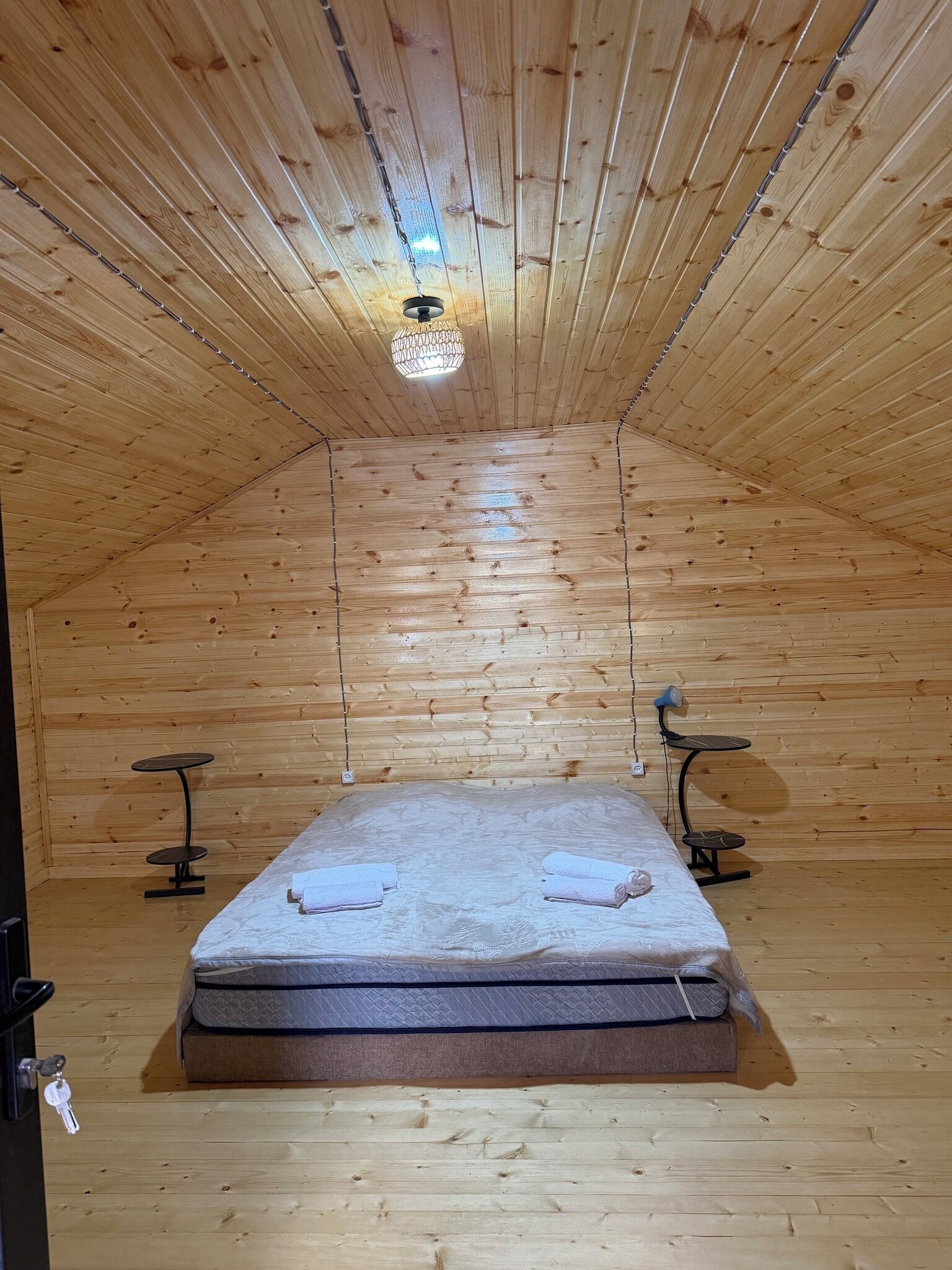 A wooden room with a bed, two small tables, a hanging light fixture, and a door with keys.