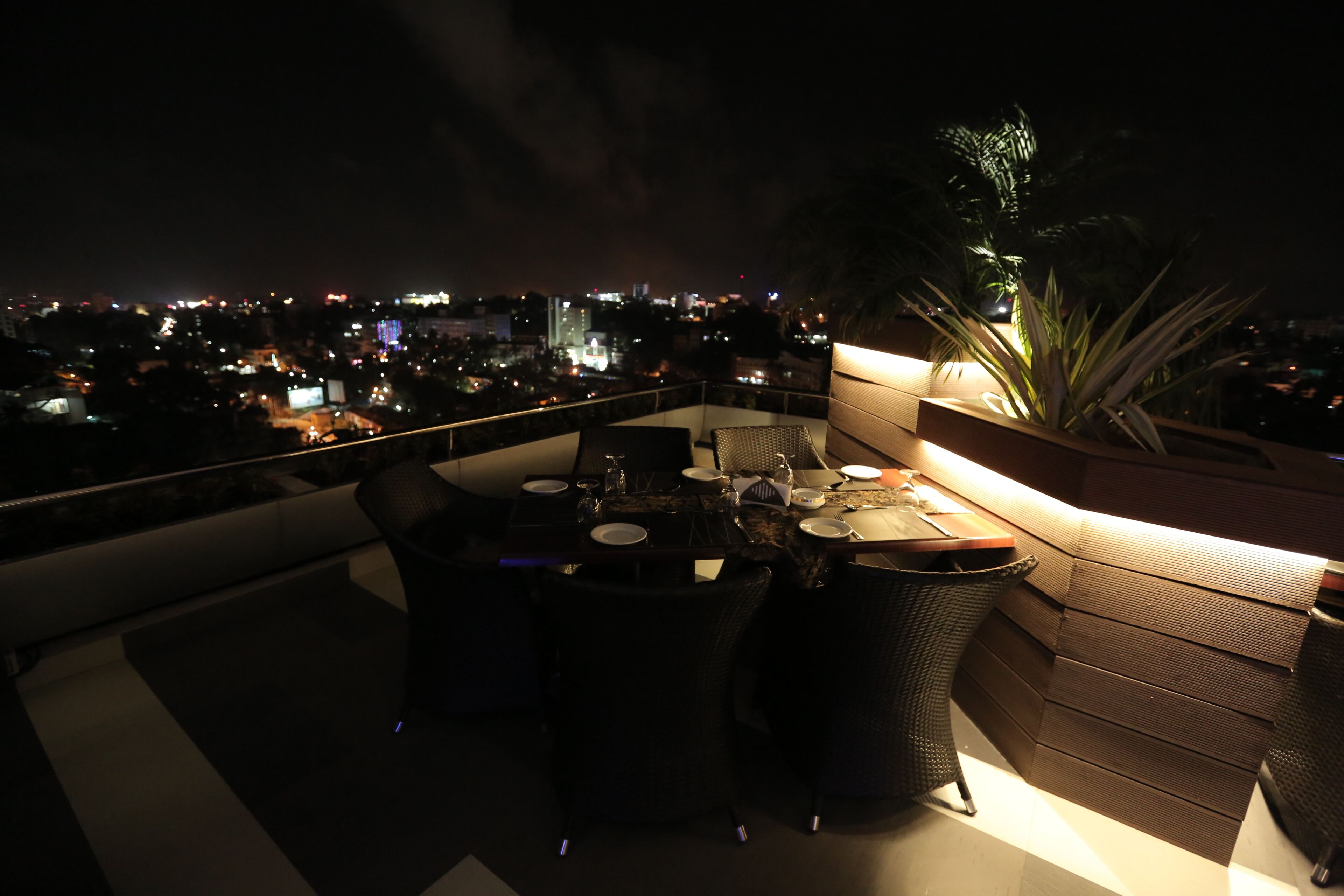Un espace de restauration aménagé sur le toit, avec une table dressée pour quatre personnes, entourée de chaises en osier, offrant une vue sur le paysage urbain illuminé la nuit.