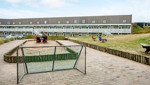A playground with a netted soccer goal, a wooden fence, and a building with multiple windows in the background. - Sondervig Holiday Center (Ringkobing)