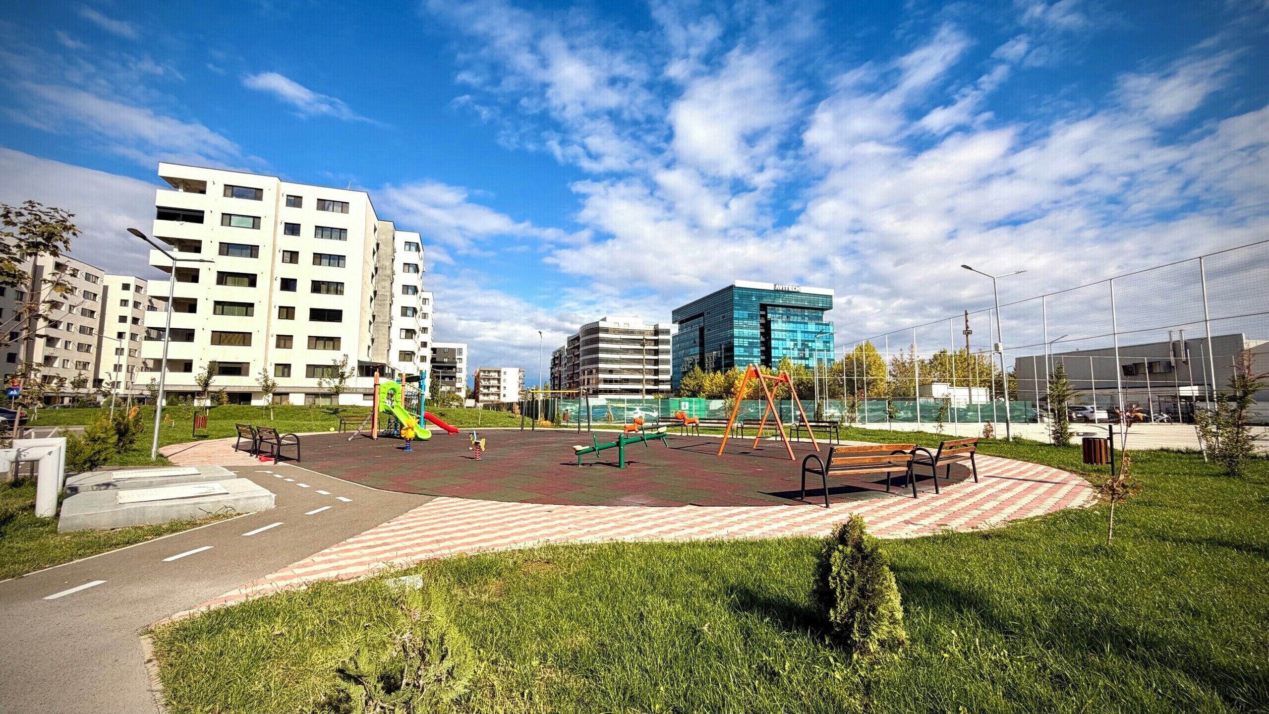 A playground with swings and a slide, surrounded by grass and a paved area.