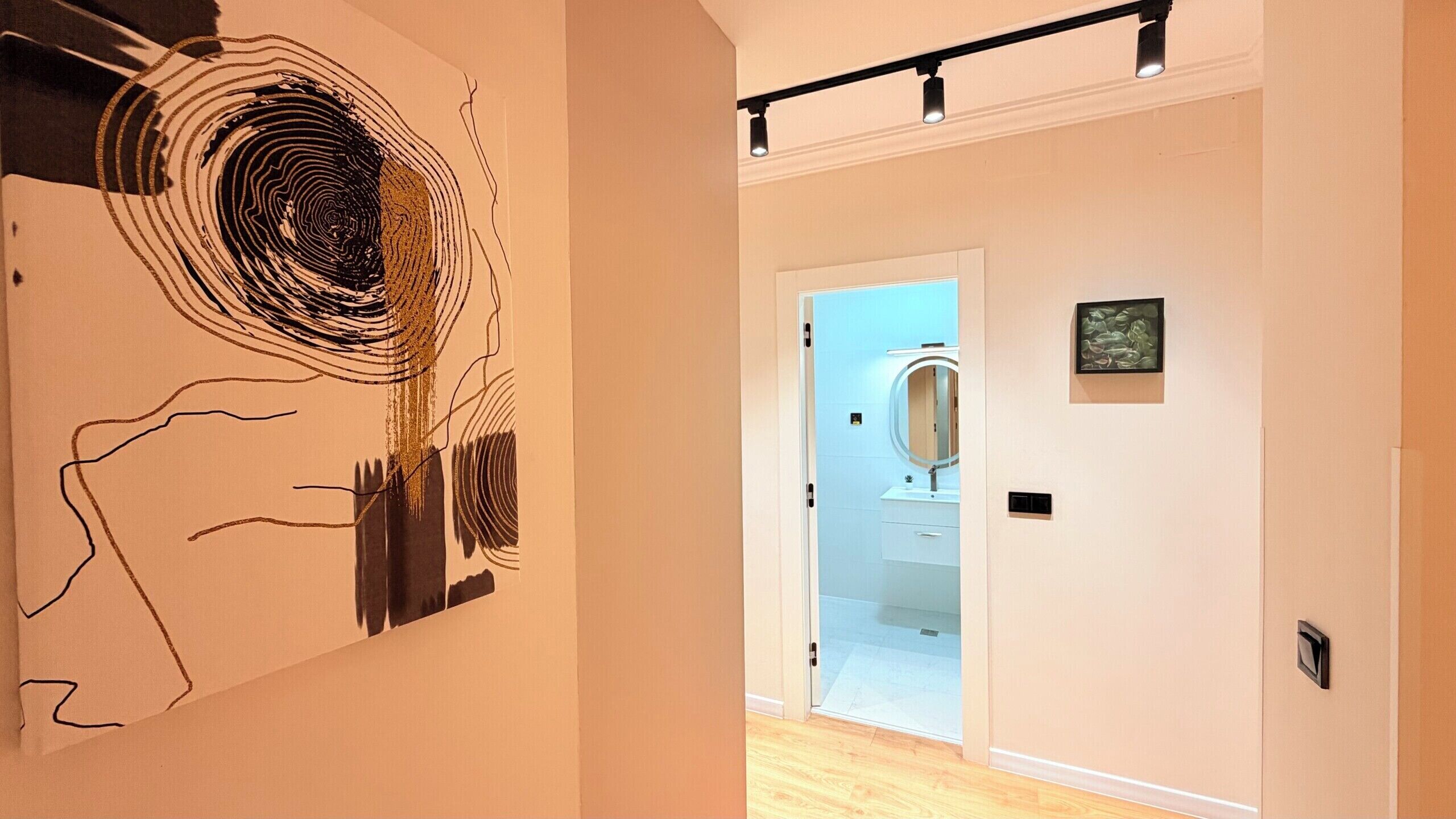 En gang med en moderne kunstinstallation til venstre, et badeværelse med spejl og håndvask til højre, og en lampe over.