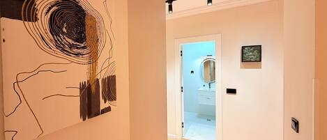 En gang med en moderne kunstinstallation til venstre, et badeværelse med spejl og håndvask til højre, og en lampe over.