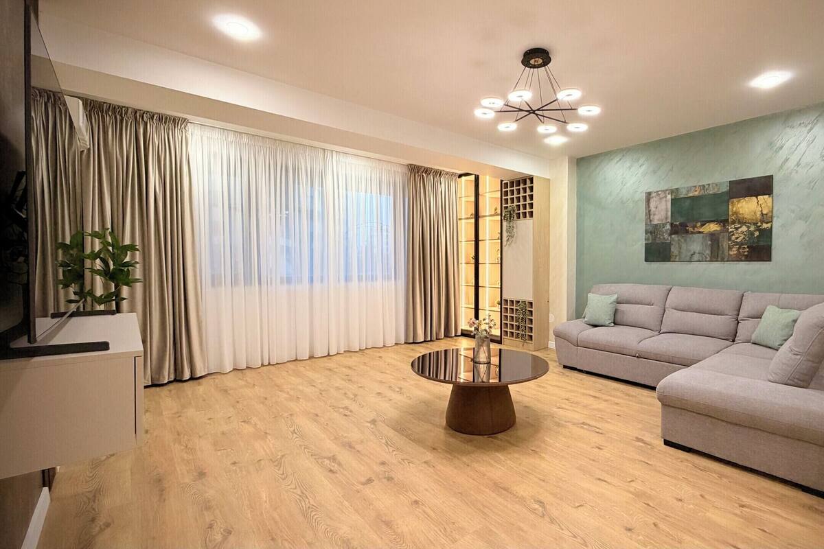 A modern living room with a round glass coffee table, a grey sofa, wooden flooring, a large window with curtains, and a ceiling light fixture.