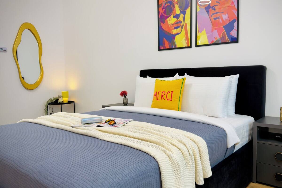 A neatly made bed with a blue and white duvet, a yellow pillow with 'MERCI' on it, and a white pillow. There is a bedside table with a lamp, a vase with flowers, and a small decorative item.
