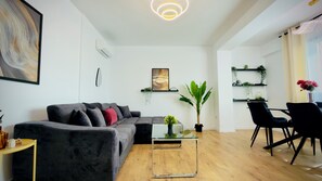 Un salon moderne avec un canapé d’angle gris, une table basse en verre, une plante en pot et un coin repas avec des chaises noires.