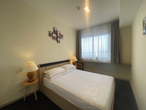 Ein Hotelzimmer mit einem Bett, zwei Nachttischlampen, einem kleinen Tisch, einem Fenster mit Vorhängen und einem an der Wand hängenden Bild.