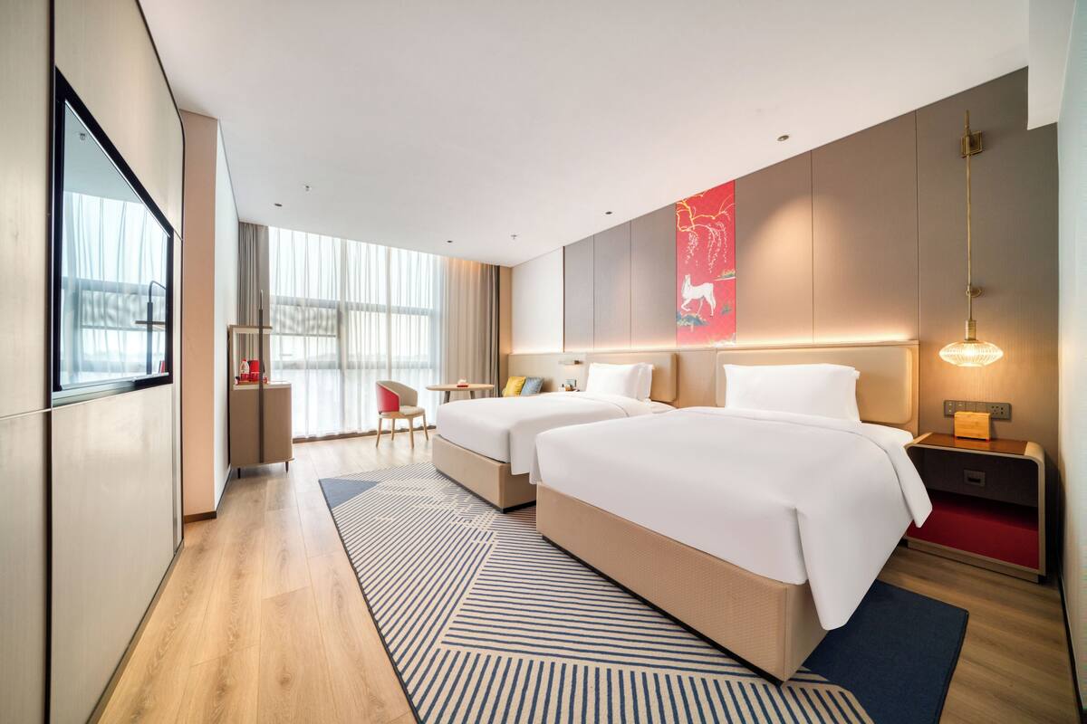 Ein modernes Hotelzimmer mit zwei Betten, einem Schreibtisch, einem Stuhl, einem großen Fenster und einem roten Kunstwerk an der Wand.