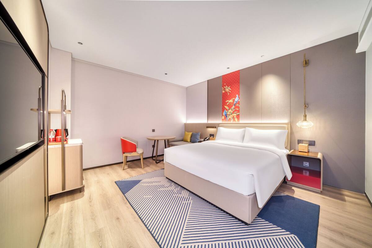 Ein modernes Hotelzimmer mit einem großen Bett, einem Schreibtisch, einem Sessel, einem kleinen Tisch, einem Fernseher und einem Nachttisch mit Lampe.