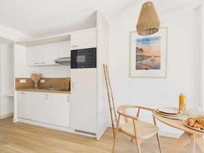 A modern kitchen with white cabinets, a microwave, and a dining area with a wooden table and chair. - Desert Point - studio for 3 - shared pool (Biscarrosse)
