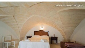 Une chambre avec un plafond en pierre, un lit recouvert de draps blancs, une table de chevet et une chaise.