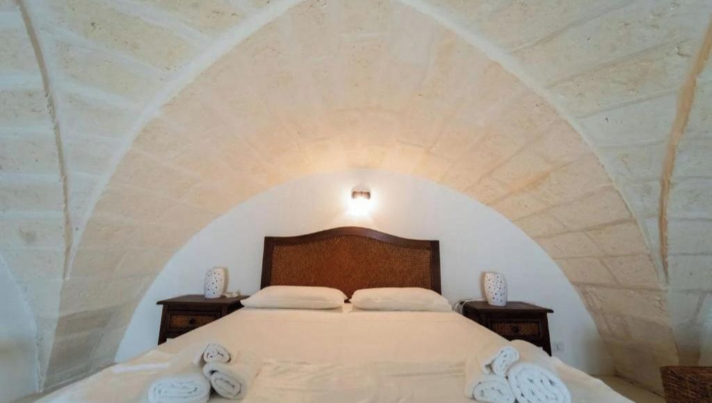 A bedroom with a stone arched ceiling, a wooden headboard, two bedside tables with decorative vases, and neatly folded towels on the bed.