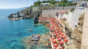 Une zone côtière avec un mur de pierres, une eau d’un bleu limpide et une rangée de parasols rouges le long du rivage.