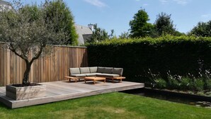 Una terraza de madera con un sofá modular, un árbol y un seto.