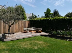 Eine Holzterrasse mit einer Eckcouch, einem Baum und einer Hecke.
