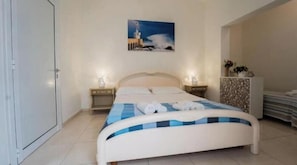 A bedroom with a bed, two bedside tables, a nightstand, a painting on the wall, and a tiled floor. - Anemone Studios - Triple room with private bathroom in Salento (Uggiano La Chiesa)