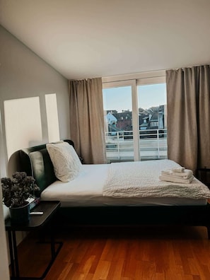 A bedroom with a bed, a bedside table, a window with curtains, and a view of buildings. - Just Sunny - Rooftop, Balcony, 3 min to Old Town (Basel)
