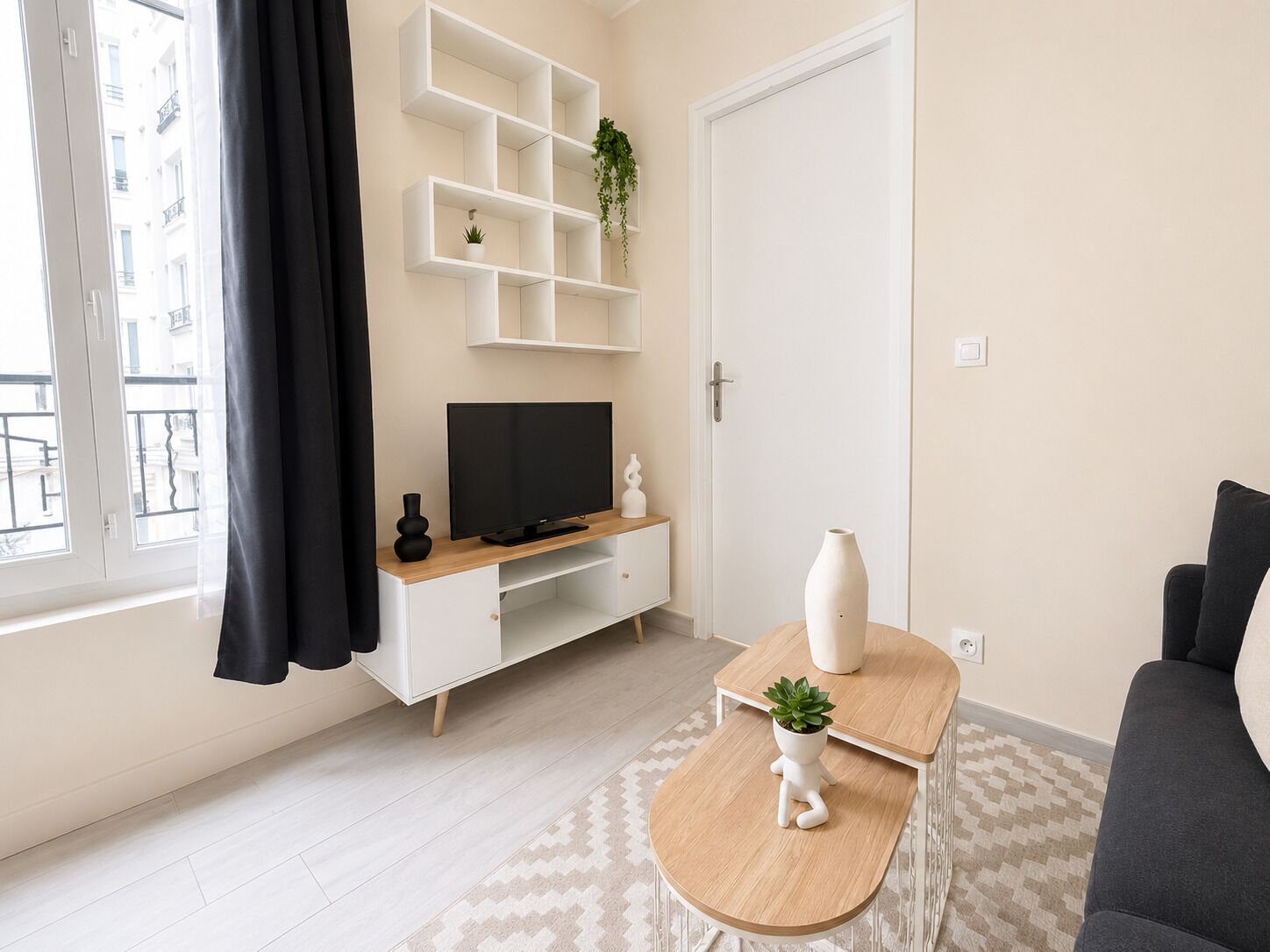 Ein modernes Wohnzimmer mit Fernseher, weißen Regalen, einem kleinen Tisch mit Pflanze und einem grauen Sofa.