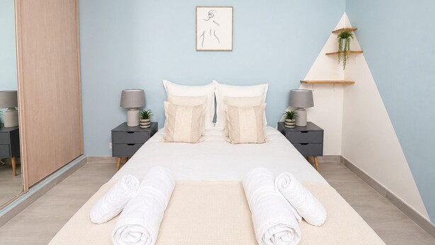 Ein Schlafzimmer mit einem Bett, zwei Nachttischen, einer Wanduhr, einem gerahmten Bild und einer Pflanze auf einem Regal.
