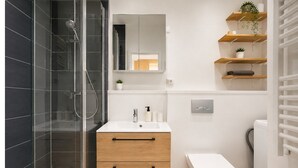 Ein modernes Badezimmer mit einer Duschkabine aus Glas, einem weißen Waschbecken mit Holzunterschrank, einer Toilette und einem an der Wand befestigten Ablagebereich mit Pflanzen.
