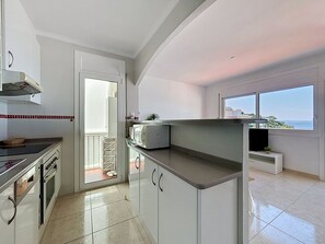 Eine moderne Küche mit weißen Schränken, einem eingebauten Backofen und einer Mikrowelle. Ein Fenster bietet Blick auf das Meer.