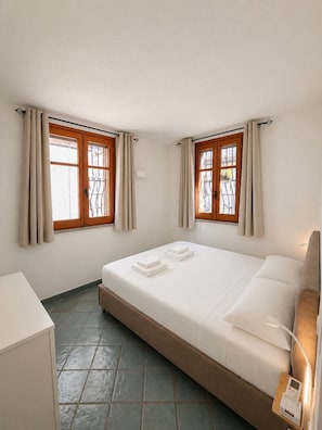 Quarto individual com uma cama e cabeceira de madeira, mesa de cabeceira com luminária e duas janelas com cortinas.