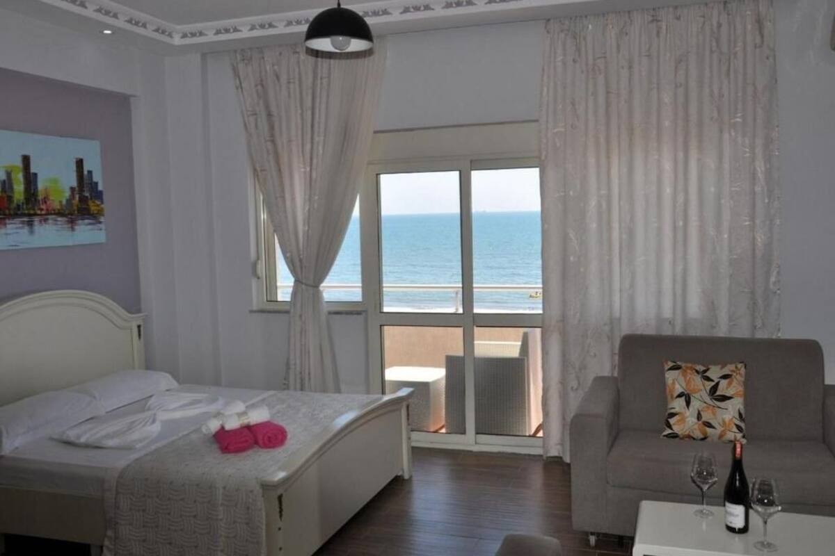 Habitación de hotel con cama, sofá, mesa de comedor con copas de vino, vista al mar y un cuadro en la pared.