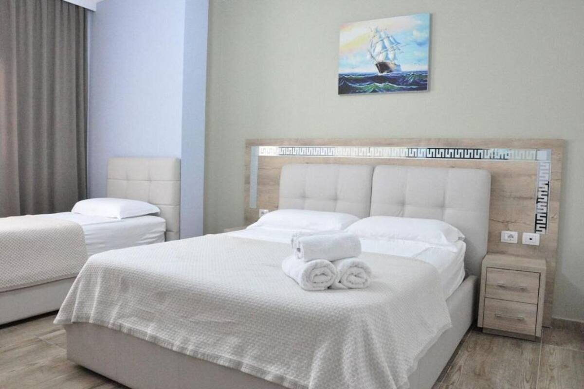 Una habitación de hotel con una cama grande, dos camas más pequeñas, un cabecero con un diseño decorativo, un cuadro de un velero y una mesita de noche con cajones.