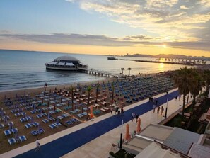 Un hotel frente a la playa con filas de tumbonas, un muelle que se adentra en el agua y una puesta de sol al fondo.
