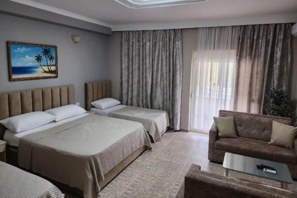 Habitación de hotel con dos camas, un sofá, una mesa de centro, un cuadro de una playa y un ventanal con cortinas.