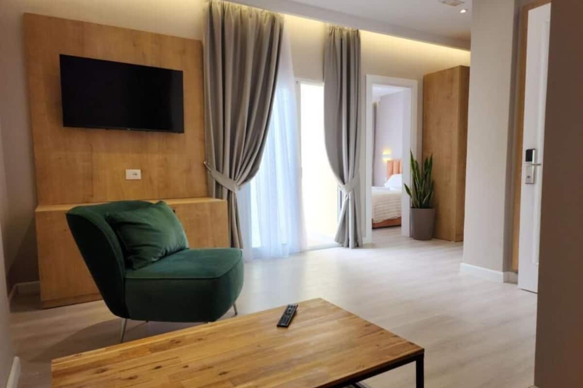 Una sala de estar moderna con un sillón verde, una mesa de centro de madera, un televisor de pantalla plana montado en una pared de madera y vista a un dormitorio a través de un vano.