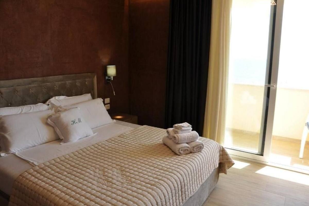 Habitación de hotel con cama, almohadas, una toalla doblada, mesita de noche, lámpara y vista al exterior a través de una puerta de vidrio.