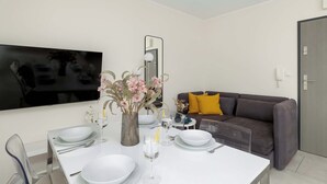 Ein modernes Wohnzimmer mit einem Esstisch für vier Personen, einem grauen Sofa mit gelben Kissen und einem an der Wand befestigten Flachbildfernseher.