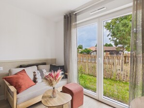 A modern living room with a sofa, a small table with a vase, and a sliding glass door leading to a garden. - Backdoor - studio for 3 - shared pool (Biscarrosse)