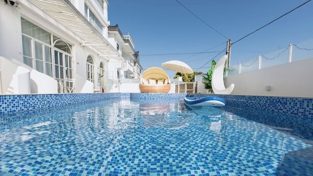 Una piscina con mosaicos azules, una silla de mimbre y un edificio blanco con ventanas arqueadas.