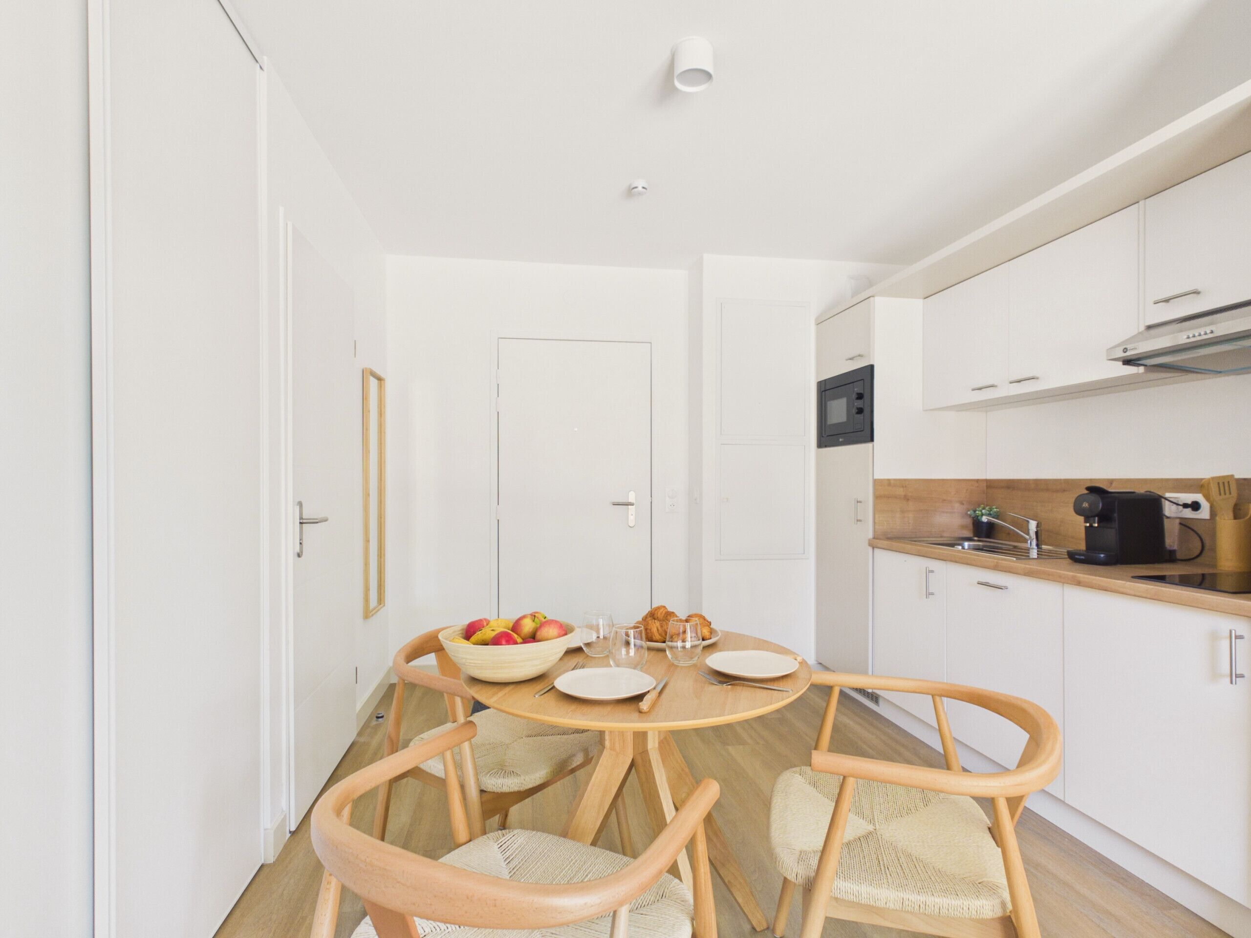 A modern kitchen with white cabinets, a wooden dining table, and a fruit bowl.