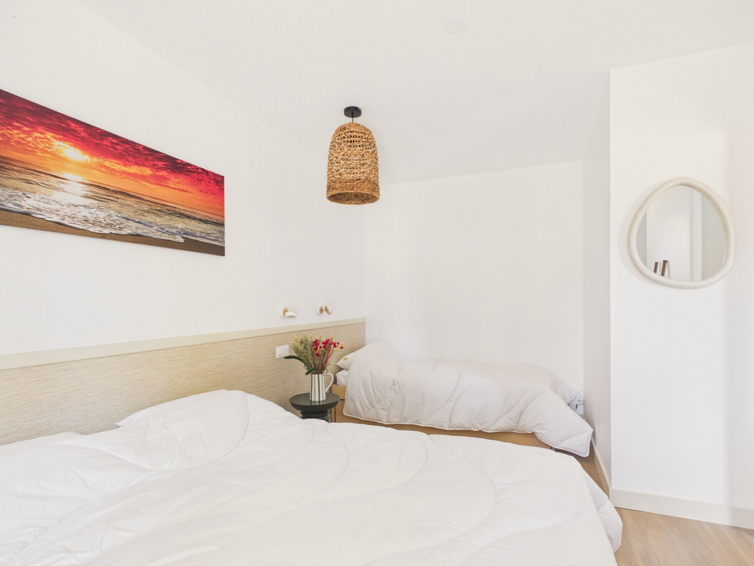 A bedroom with a bed, a bedside table with a vase of flowers, a wall-mounted lamp, and a round mirror.