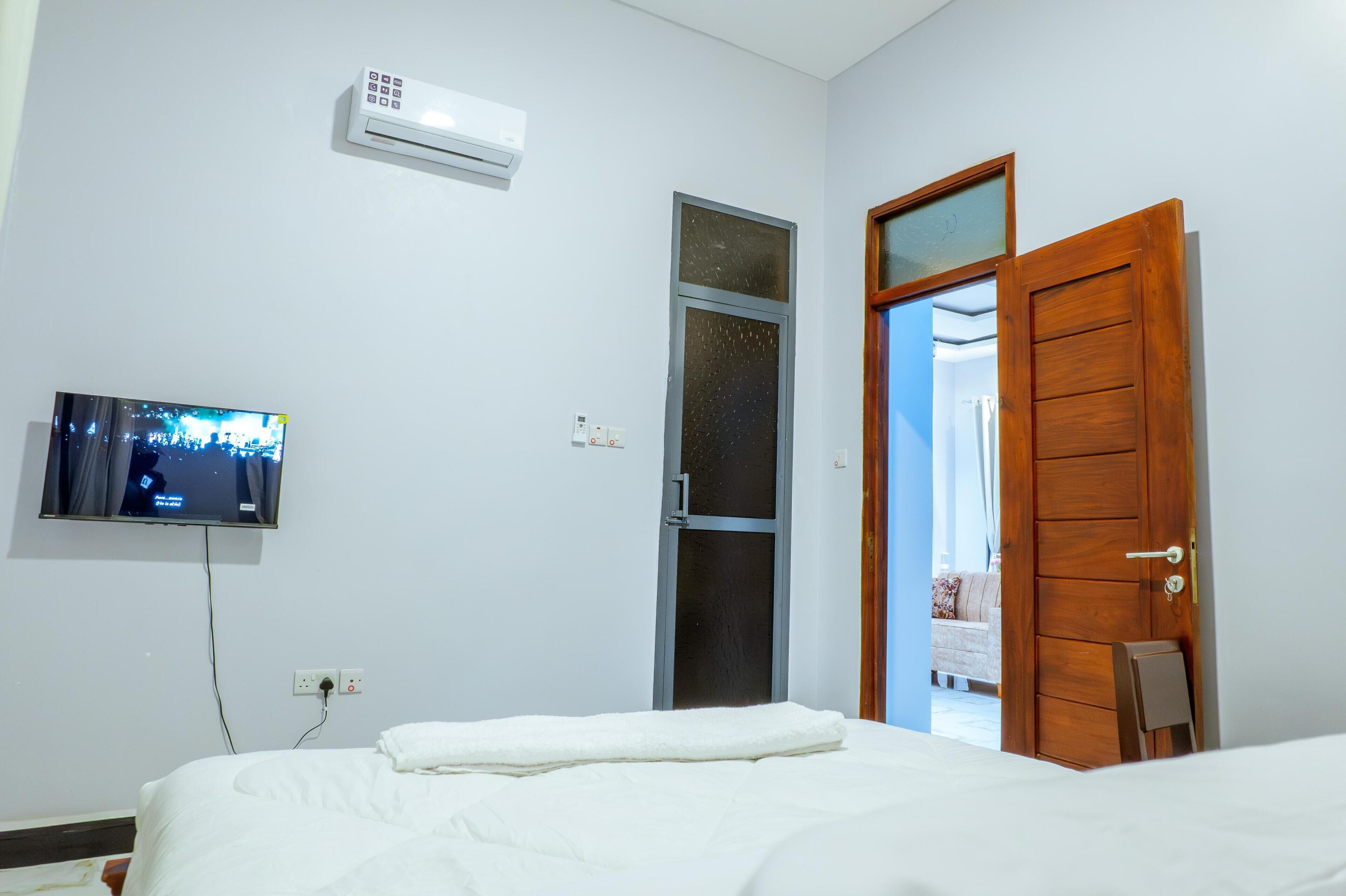 Un dormitorio con cama, televisión montada en la pared, aire acondicionado, puerta y una mesita.