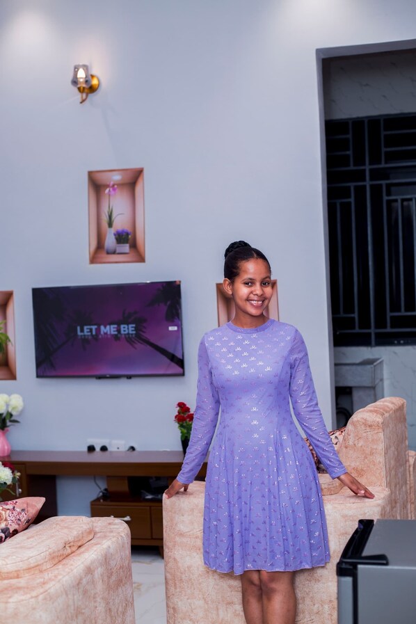 A person standing in a room with a television displaying 'LET ME BE' on the screen, a wall-mounted light fixture, and a small framed picture.