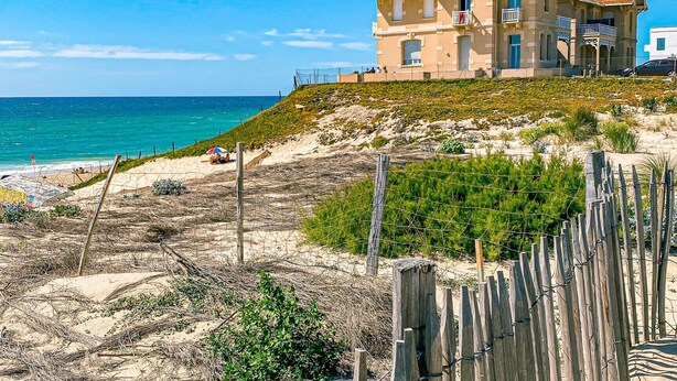 Une maison située sur la côte, avec vue sur la plage, une clôture en bois et un ciel dégagé.