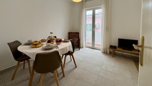 A dining area with a table set for four, chairs, a television on a stand, and a sliding glass door leading to an outdoor area. - Lagos Design 5 by Homing (Lagos)