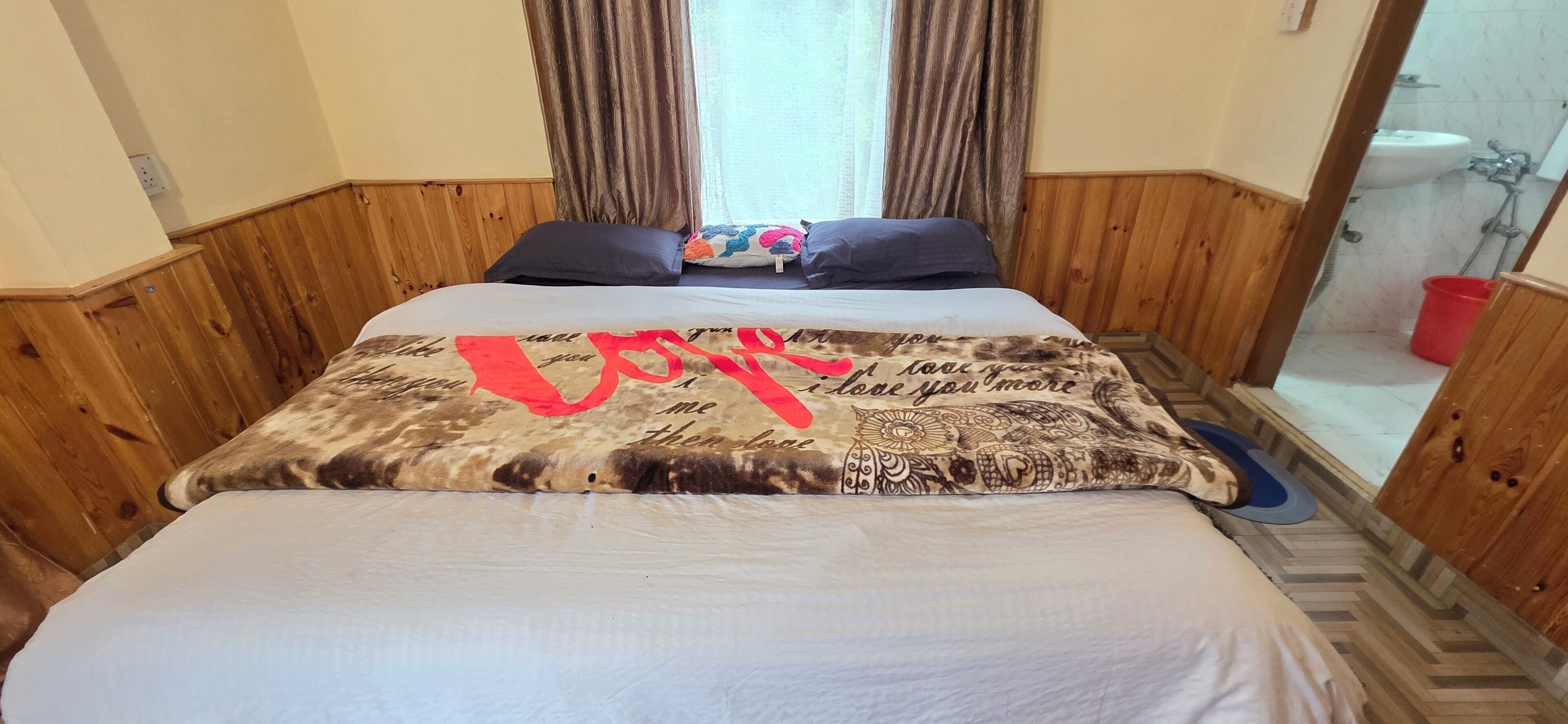 Camera d'albergo con un letto, pannelli in legno, una finestra con tende e un bagno visibile attraverso una porta aperta.