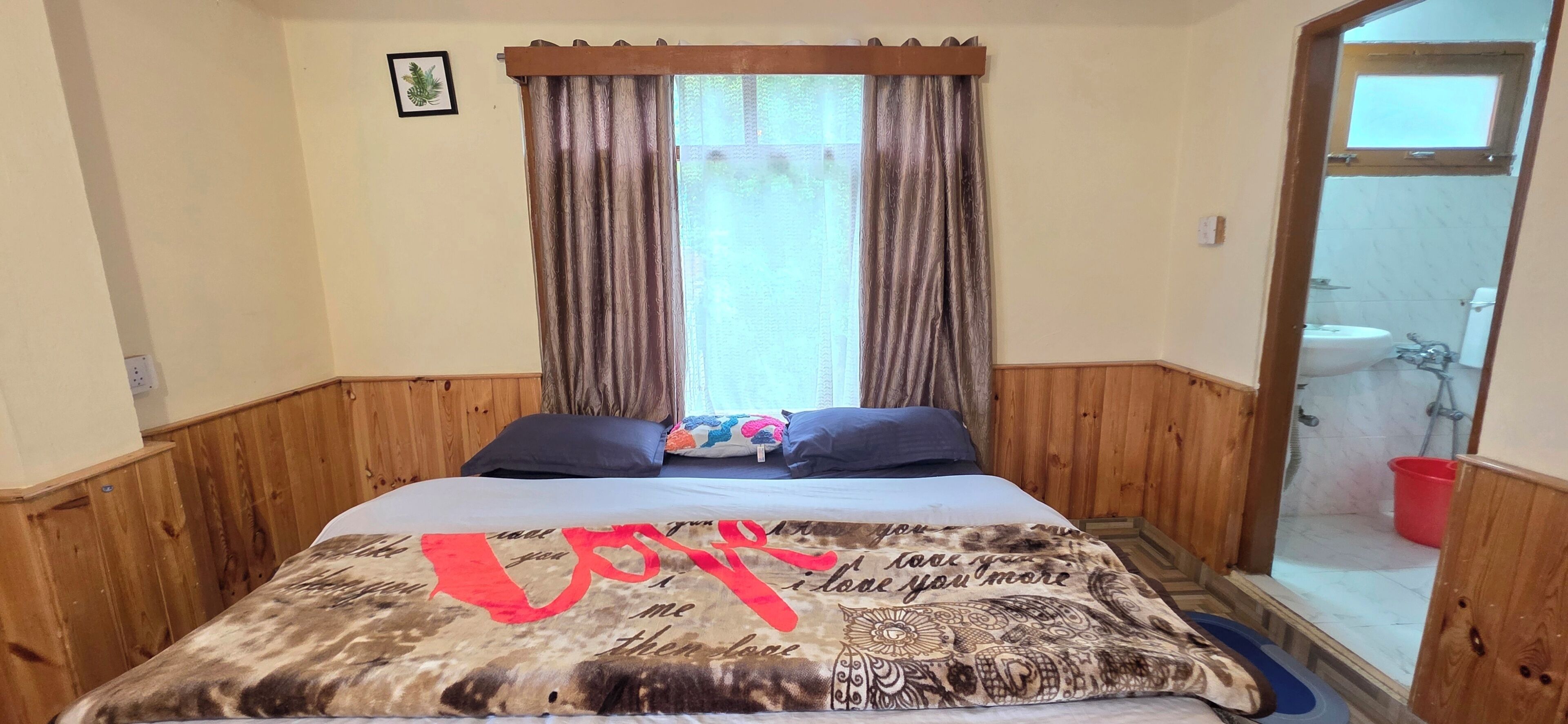 A bedroom with a bed, wooden paneling, a window with curtains, a bathroom visible through a glass door, and a small framed picture on the wall.