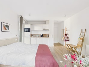 A bedroom with a bed, a red blanket, a kitchenette, and a wooden chair. - Uluwatu – studio for 2 – shared pool (Biscarrosse)