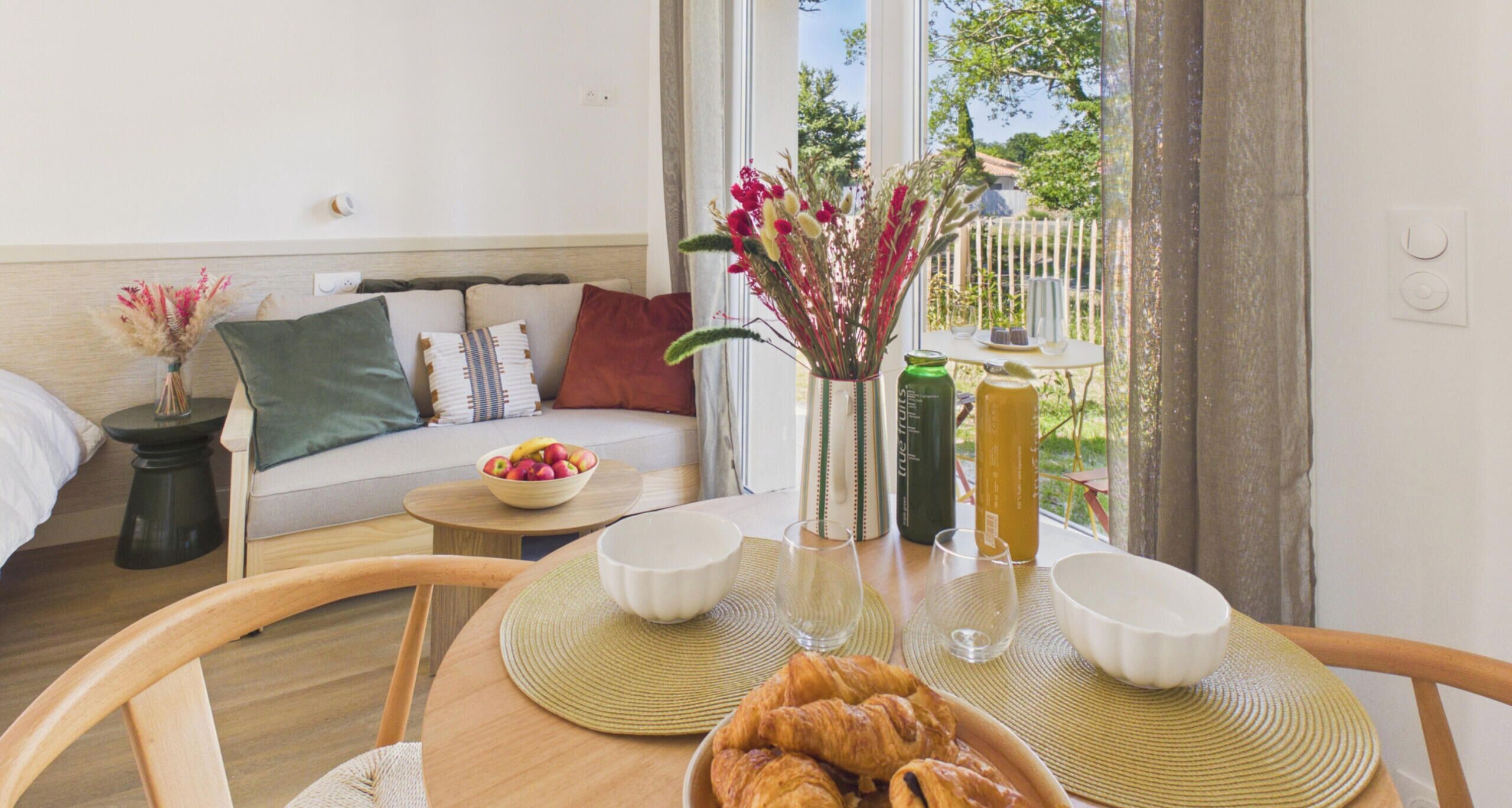 A cozy room with a sofa, a dining table with croissants, a bowl of fruit, and a vase with flowers.