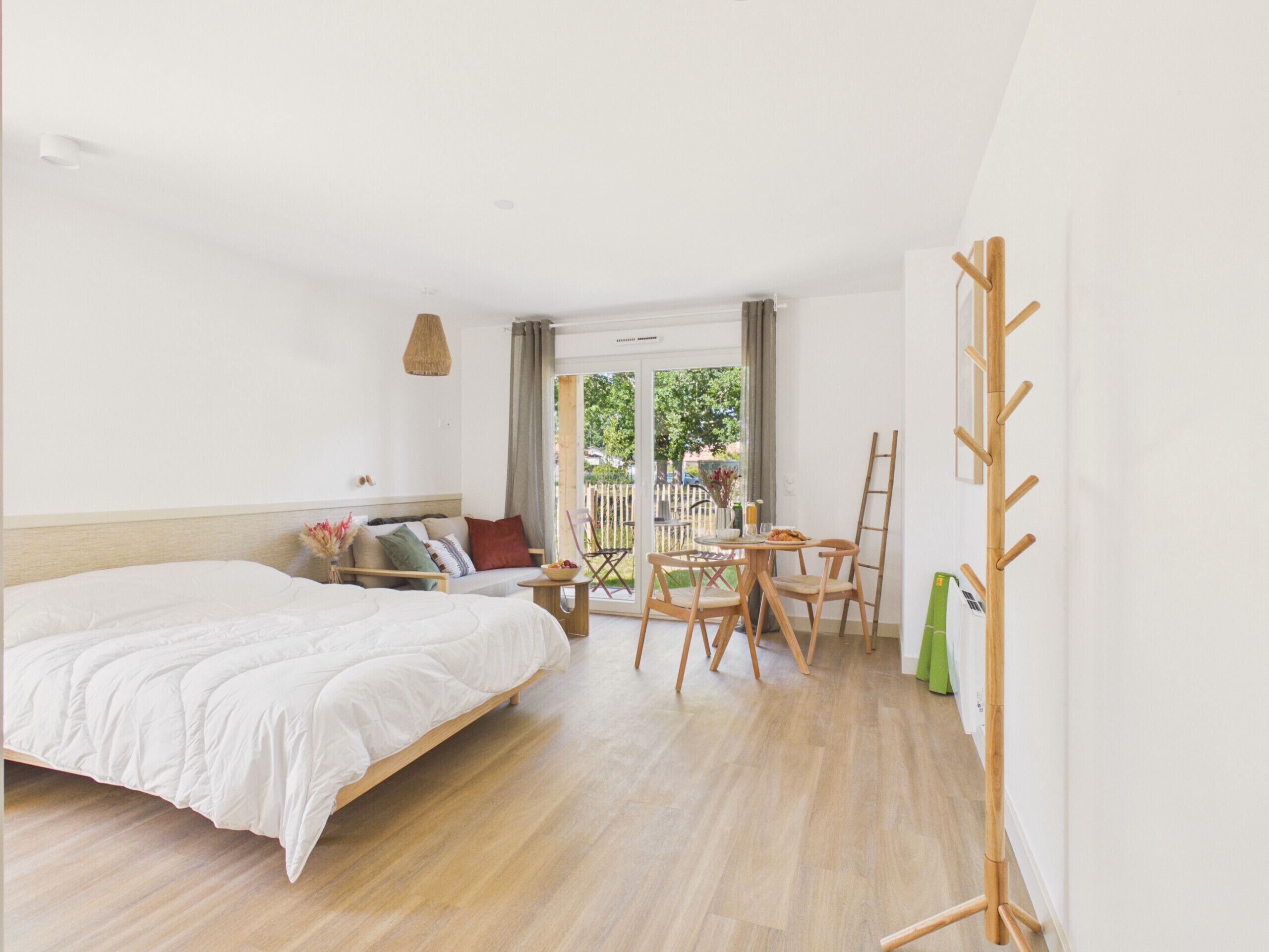 A bedroom with a bed, a small table with chairs, a wooden ladder, and a view of the outdoors.