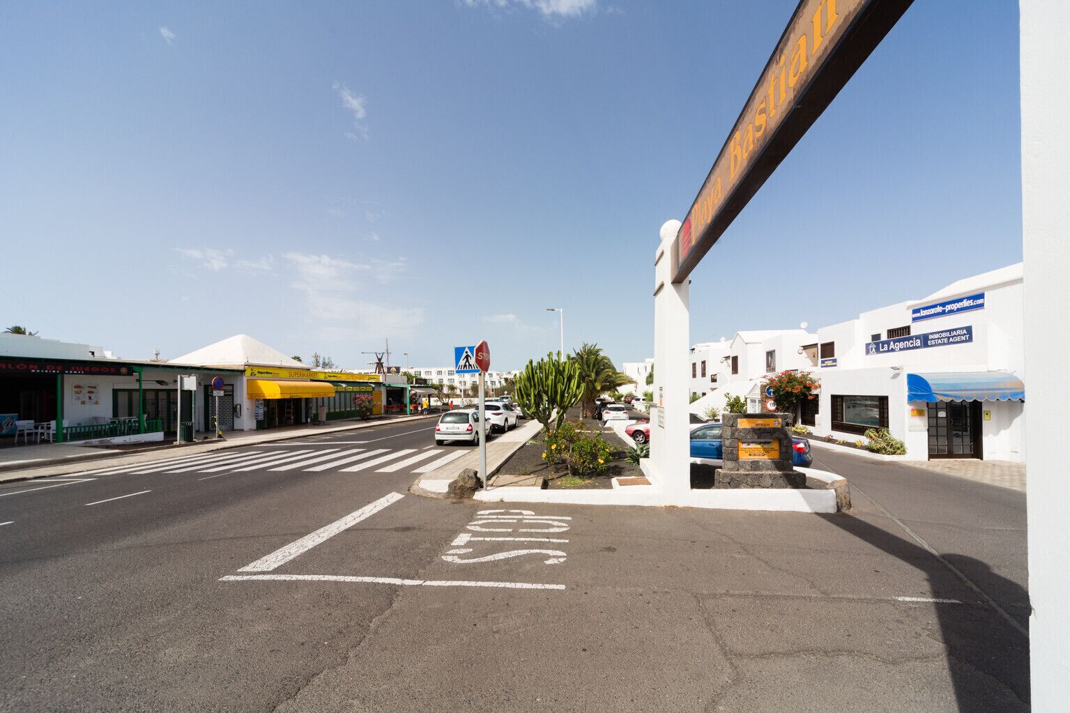 Vista da rua com faixa de pedestres, placa de pare e edifícios, incluindo um supermercado e uma imobiliária.