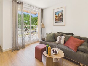 Een moderne woonkamer met een bank, een klein rond tafeltje met een fles en glazen, en een balkon met uitzicht op groen.