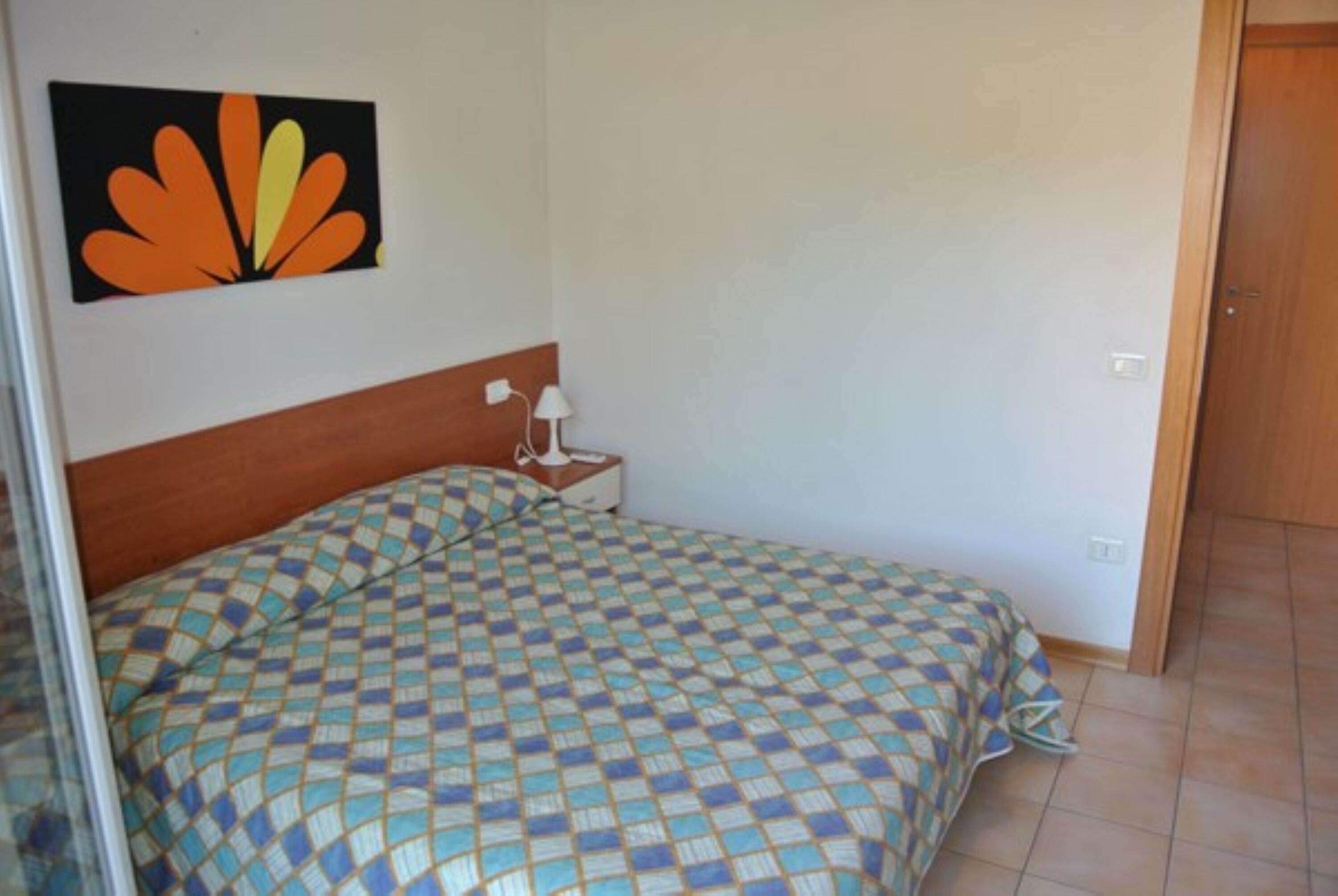 A bedroom with a checkered bedspread, a nightstand with a lamp, and a wall-mounted picture.