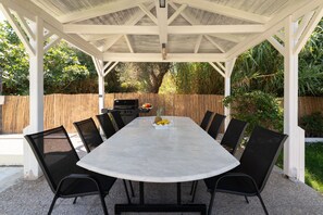 Eine überdachte Terrasse im Freien mit einem rechteckigen Tisch und Stühlen, einem Grill und einem Holzzaun.