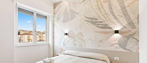 Une chambre d’hôtel avec un lit, une fenêtre, un mur au décor texturé et une table de chevet avec une lampe.